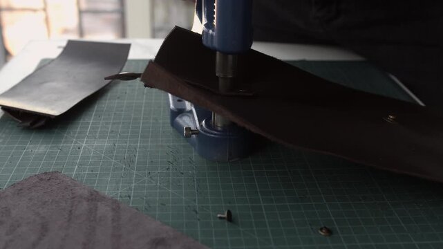 Leather craftsman using a manual press to set rivets on a dark brown strap in a traditional workshop with a cutting mat