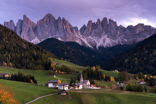 View of the St. Magdalena church nestled in green meadows with the dramatic jagged peaks of the Odle Mountains in the background under a soft evening sky in Italy.