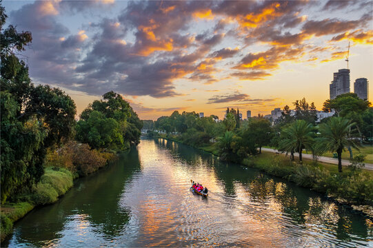 Aerial view of Yarkon River with a boat carrying people, lush greenery, and the city skyline under a vibrant sunset sky in Tel Aviv-Yafo, Tel Aviv District, Israel.