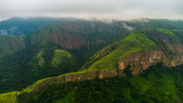 Aerial view of the lush green mountains and rocky cliffs of Likpe Bakwa with a distant waterfall under a cloudy sky in Likpe Bakwa, Volta Region, Ghana.