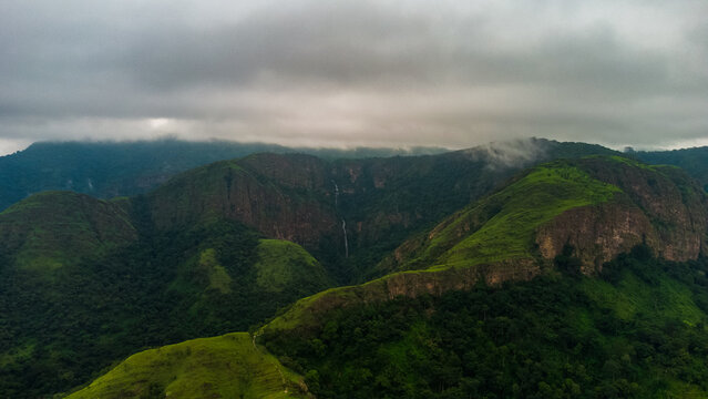 Aerial view of the lush green mountains and cliffs of Likpe Bakwa with a distant waterfall under a cloudy sky in Likpe Bakwa, Volta Region, Ghana.