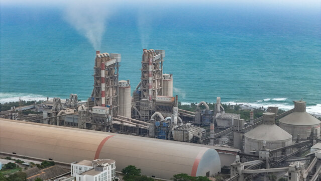 Aerial view of a large industrial cement factory with tall processing towers emitting smoke and storage silos situated along the coastline by the sea in Indonesia.