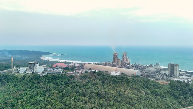 Aerial view of a large industrial cement factory situated on the coast, surrounded by lush green forest with the ocean in the background, Indonesia.