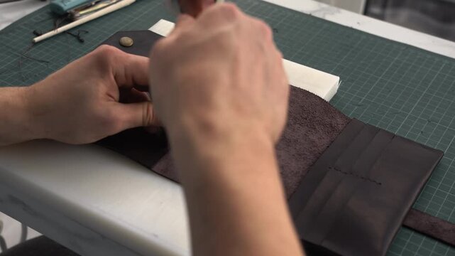 Craftsman punching stitching holes in brown leather wallet using chisel and maul in a workshop