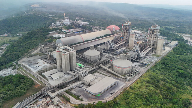 Aerial view of a large industrial cement factory complex with silos and processing buildings situated in a forested hilly landscape in Indonesia.