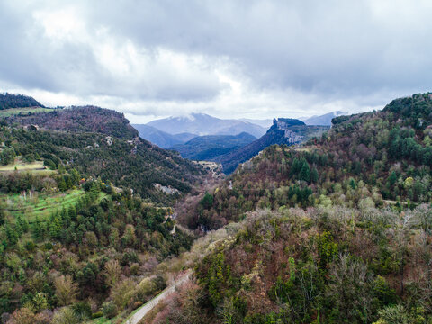 Aerial view of the rugged mountainous landscape featuring dense forests, rocky cliffs, and a winding road under a cloudy sky in Rupit, Catalonia, Spain.