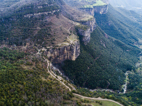 Aerial view of the rugged cliffs and dense forests surrounding Mas el Roquer, featuring winding dirt paths and dramatic rock formations in Rupit, Catalunya, Spain.