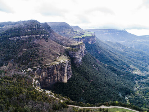 Aerial view of Mas el Roquer surrounded by dramatic rocky cliffs, winding trails, and dense forest under a cloudy sky in Rupit, Catalunya, Spain.