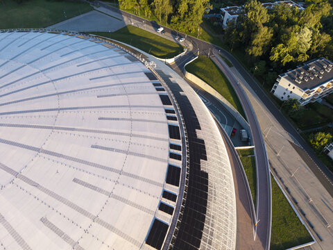 Aerial view of the large domed roof of Tauron Arena Krakow featuring integrated solar panels, surrounded by green spaces and urban roads in Krakow, Lesser Poland Voivode, Poland.