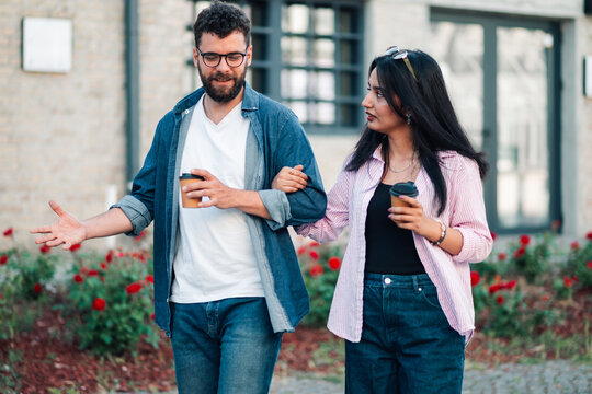 Young couple walking arm in arm, holding coffee cups, having a conversation during an urban date