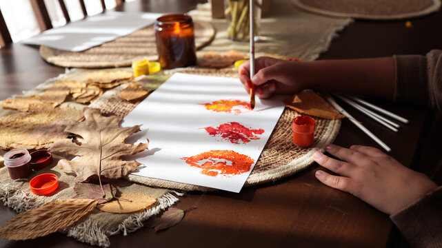 Child's hands painting autumn leaf prints with vibrant colors on white paper