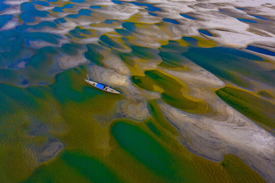 Bogura, Bangladesh - 03 January 2022: Aerial view of a traditional wooden boat navigating the shallow, patterned sandbars and vibrant green waters of the Jamuna River near Sariakandi.