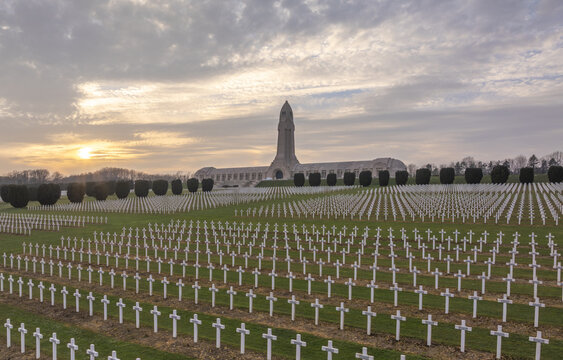 Aerial view of the Douaumont Ossuary and the surrounding military cemetery with rows of white crosses on green grass under a soft sunset sky in Douaumont-Vaux, Grand Est, France.