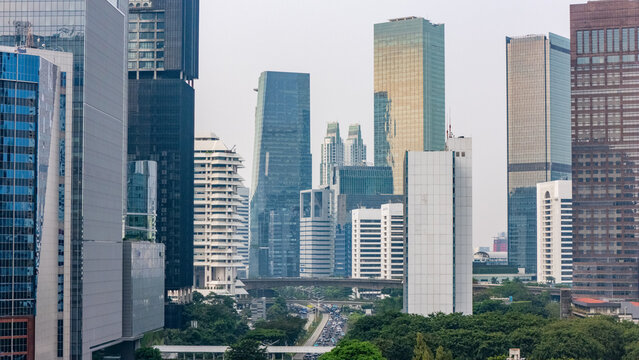 Aerial view of modern skyscrapers and office buildings rising above lush green trees and urban infrastructure in the city center of Jakarta, Indonesia.