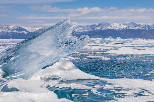 View of a large, jagged, transparent blue ice shard on the frozen surface of Lake Baikal with snow-covered mountains in the background under a clear blue sky in Khuzhir, Irkutsk Oblast, Russia.
