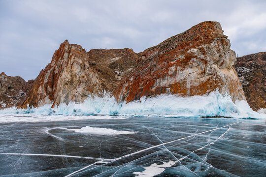 View of the frozen surface of Lake Baikal with dramatic orange and brown rocky cliffs and snow-covered base under an overcast sky, Khuzhir, Irkutsk Oblast, Russia.