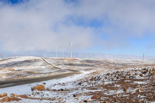 View of wind turbines in a snowy landscape with a winding road in Al-Qadisiya, Tafilah Governorate, Jordan.