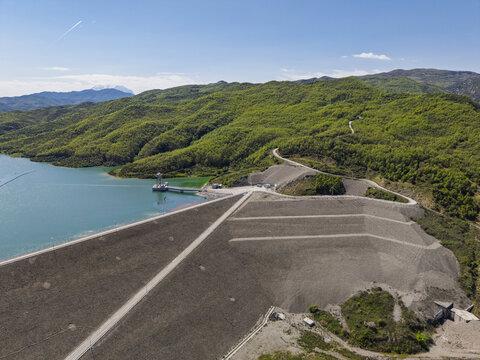 Aerial view of Shushice Dam and reservoir surrounded by lush green hills under a clear blue sky in Shushice, Elbasan County, Albania.