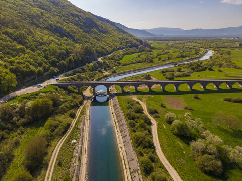 Aerial view of Tsar's Bridge spanning the Zeta River in a lush green valley with rolling hills, clear blue water, and a bright sunny sky in Niksic, Opstina Niksic, Montenegro.