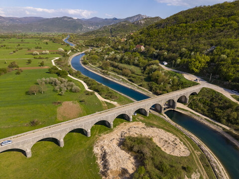Aerial view of the historic stone Tsar's Bridge spanning a river through lush green fields and rolling hills in Niksic, Opstina Niksic, Montenegro.
