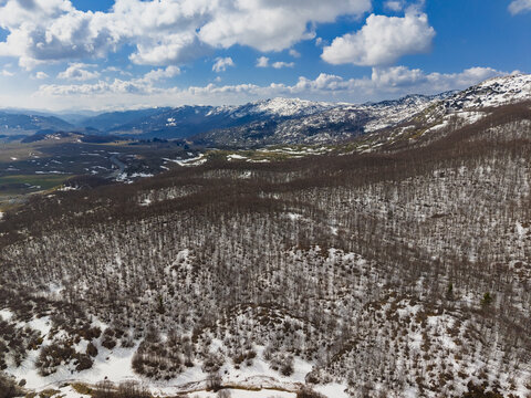 Aerial view of the snow-covered slopes and dense forests of Durmitor National Park under a bright blue sky with scattered clouds in Zabljak, Zabljak Municipality, Montenegro.