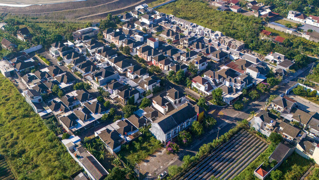 Aerial view of a residential neighborhood with rows of houses featuring dark roofs, surrounded by lush greenery and agricultural plots in Indonesia.