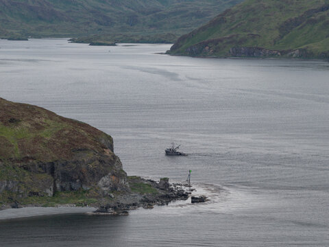 Aerial view of a small fishing boat navigating the calm waters of the bay near the rocky coastline of Unalaska, Alaska, United States.