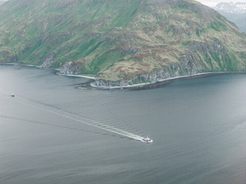 Aerial view of a boat cruising through the deep waters of a bay near the rugged green mountains of Unalaska, Alaska, United States.