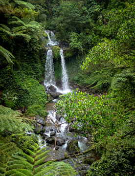 Aerial view of the lush Grenjengan Kembar Waterfall cascading down into a rocky stream surrounded by dense tropical rainforest and ferns in Kecamatan Pakis, Jawa Tengah, Indonesia.