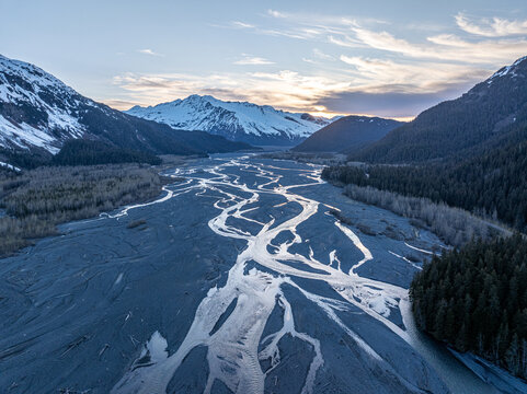Aerial view of the braided Resurrection River flowing through a valley surrounded by snow-capped mountains and evergreen forests under a soft sunset sky in Seward, Alaska, United States.