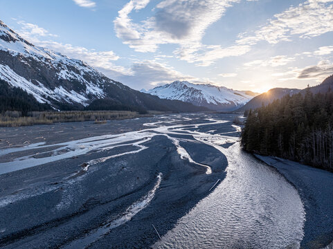 Aerial view of Resurrection River flowing through a braided riverbed surrounded by snow-capped mountains and evergreen forests under a golden sunset in Seward, Alaska, United States.