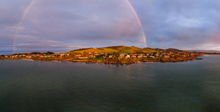 Aerial view of Riverton with a rainbow arching over the coastal town, green hills, houses, and calm ocean waters in Riverton, Southland Region, New Zealand.