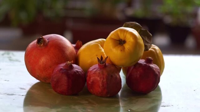 Slow motion camera panning of fresh red pomegranates and ripe yellow quinces on a glossy table with a blurred garden background, horizontal video.