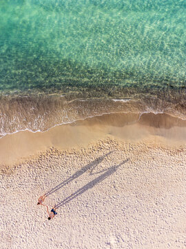Aerial view of a couple holding hands on a sandy beach, their long shadows stretching across the sand beside turquoise water during golden hour in Sardinia, Italy.
