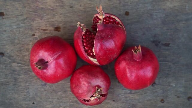 Top view of ripe red pomegranates with one cracked open revealing juicy seeds on a rustic wooden table, slow motion horizontal video.
