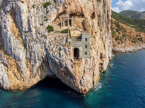 Aerial view of the historic Porto Flavia mining site built into the sheer limestone cliffs overlooking the turquoise Mediterranean Sea in Sardinia, Italy.