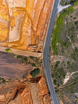 Aerial view of the abandoned Monteponi mine, its layered orange and brown soil textures bordering a winding asphalt road and green vegetation in Sardinia, Italy.