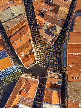 Aerial view of a street decorated with colorful umbrellas suspended between historic buildings with terracotta rooftops in Sardinia, Italy.