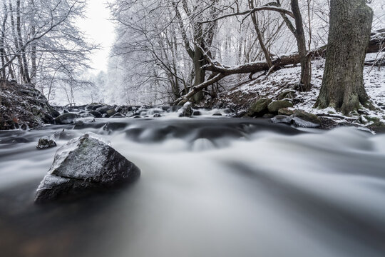 View of a flowing stream with long-exposure water, ice-covered rocks, and snow-dusted trees in a winter forest in Debrzno, Pomeranian Voivodeship, Poland.