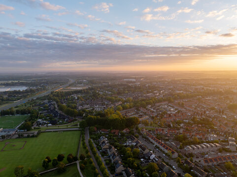 Aerial view of a residential neighborhood in the Netherlands at sunset, featuring rows of houses, green sports fields, and tree-lined streets under a warm golden sky.