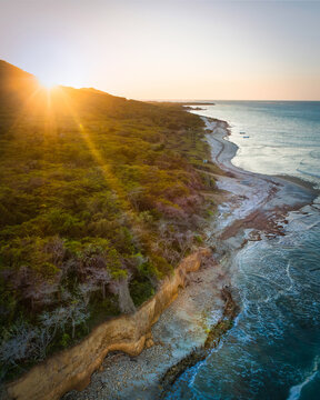 Aerial view of Playa Popa coastline at sunset, with golden sunlight hitting lush forest, rugged cliffs, and the Caribbean Sea in Los Manatiales, Monte Cristi, Dominican Republic.