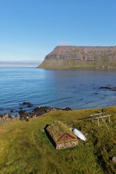 Aerial view of a traditional turf-roofed Viking hovel on a grassy coastline near the sea, with a small boat and dramatic mountain in Sudureyri, Isafjardarbaer, Iceland.