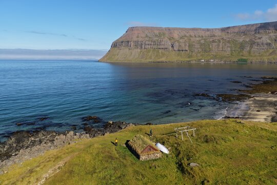Aerial view of a traditional turf-roofed Viking hovel situated on a grassy coastal cliff overlooking the calm blue sea and rugged mountains in Sudureyri, Isafjardarbaer, Iceland.