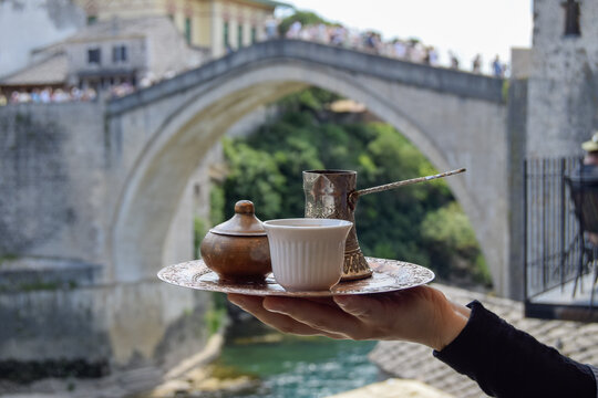 View of a traditional Bosnian coffee set with a copper pot and white cup on a tray, held in front of the historic Stari Most bridge in Mostar, Federation of Bosnia, Bosnia and Herzegovina.