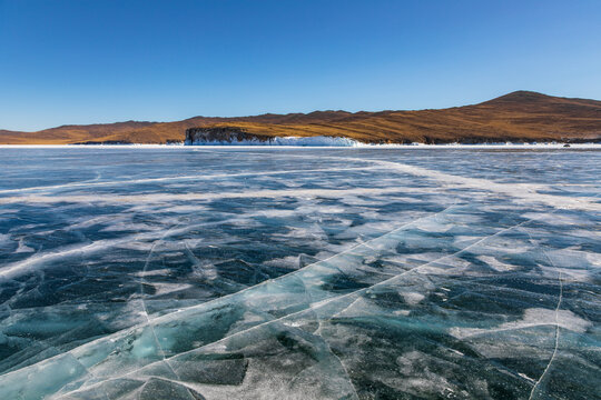 View of the frozen surface of Lake Baikal with intricate ice cracks and patterns under a clear blue sky, with rolling hills in the background in Khuzhir, Irkutsk Oblast, Russia.