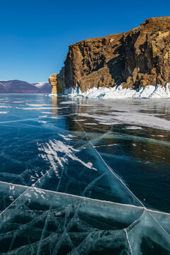 View of the frozen surface of Lake Baikal with intricate ice cracks and towering rocky cliffs under a clear blue sky in Khuzhir, Irkutsk Oblast, Russia.