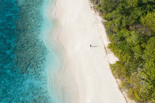 Aerial view of a person walking along the white sand beach bordered by lush green tropical vegetation and vibrant turquoise coral reef waters in Muraidhoo, Haa Alif Atoll, Maldives.