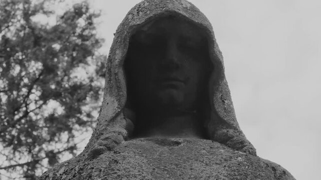 Eerie, disfigured stone statue in a cemetery, shrouded in shadow with a gray, overcast sky and dark trees in the background. Creates a somber and mysterious atmosphere.
