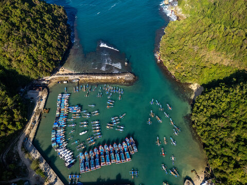 Aerial view of Pantai Sadeng with numerous colorful fishing boats moored in the turquoise bay surrounded by lush green hills in Girisubo, Special Region of Yogyakarta, Indonesia.