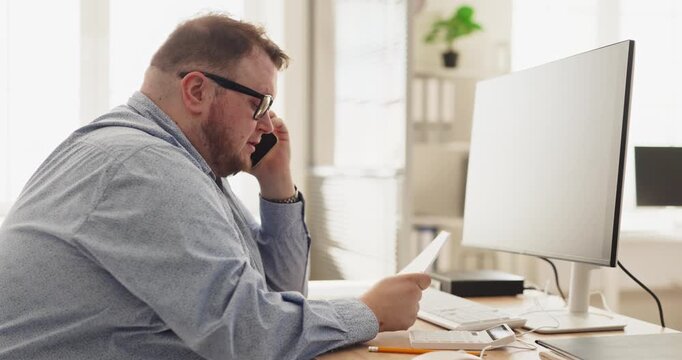 Man on phone talking about bill payment. Seated at a computer in a home office, he reviews paperwork, checks budget, and calls support to resolve charges. Theme is personal finance and bills.